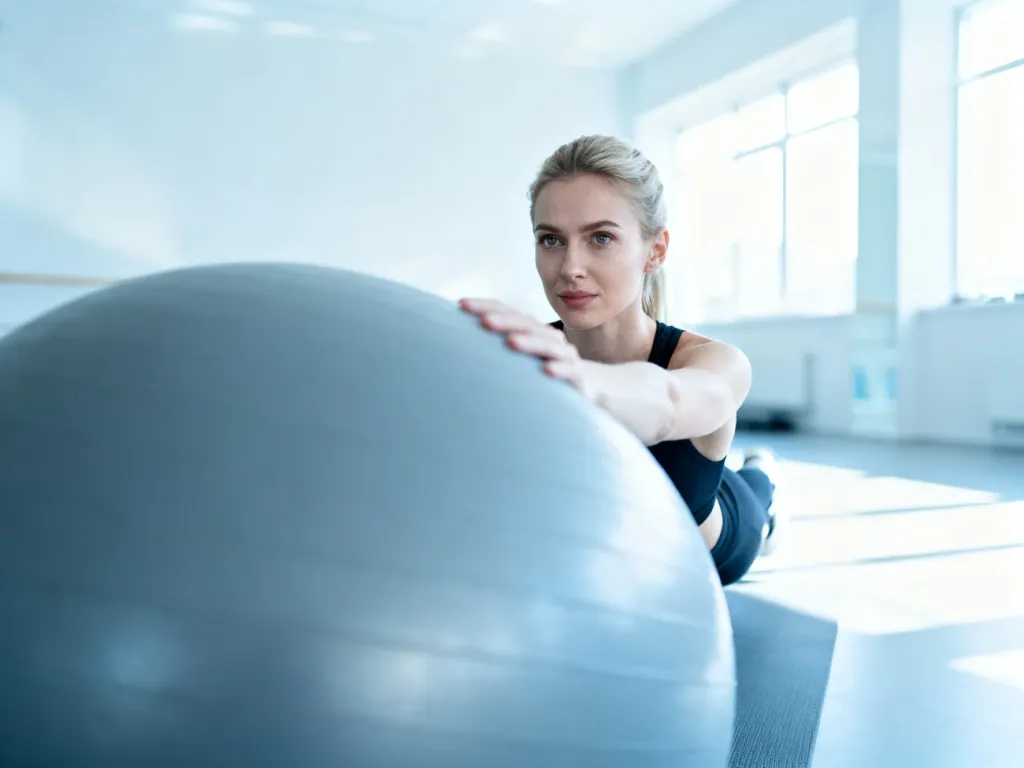 Kobieta wykonuje ćwiczenia wzmacniające mięśnie głębokie na szarej piłce gimnastycznej w nowoczesnym studio fitness.